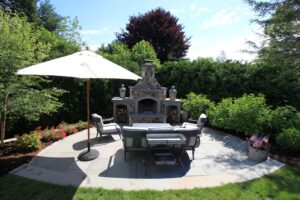 Covered patio with stone fireplace, mounted TV, wood furniture, and shaded seating surrounded by trees.