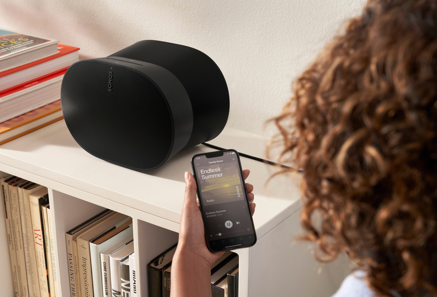 A person using the Sonos app to control music on a black Sonos speaker placed on a bookshelf filled with books and decor items.